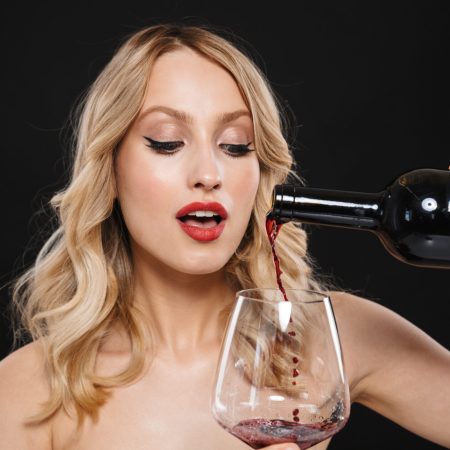 Young blonde woman with bright makeup red lips posing isolated over black wall background drinking red wine. Portrait of a pretty young blonde woman with bright makeup red lips posing isolated over black wall background drinking red wine.